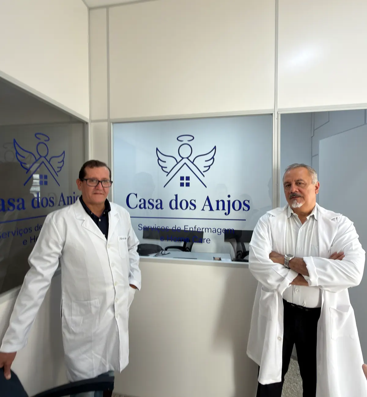 Flávio e Daniel, sócios da Casa dos Anjos, de pé na recepção da sede, com o logo no vidro entre os dois